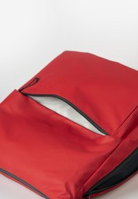 Roter Rucksack mit einer glatten, matten Oberfläche und einem Reißverschluss-Eingang. Innen mit grauem Stoff ausgekleidet, mit einem schwarzen Reißverschluss-Detail.