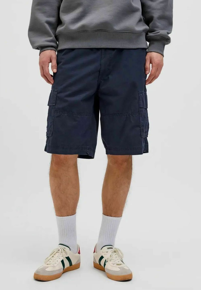 Persona vistiendo pantalones cortos cargo azul marino, calcetines blancos hasta la mitad de la pantorrilla, zapatillas retro blancas con rayas verdes y una sudadera gris.