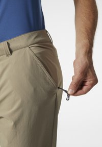 Beige broek met een gladde textuur heeft een zijzak, waaruit een zwarte koord wordt getrokken. De tailleband is iets zichtbaar.