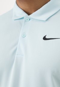 Jasnoniebieska koszulka polo o miękkiej fakturze, z czarnym logo Nike i dwoma jasnoniebieskimi guzikami na kołnierzu.