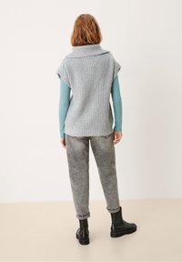 Ljusgrå stickad ärmlös tröja med upprullad krage, lagd över en blå långärmad skjorta, parat med blekta grå jeans och svarta stövlar.