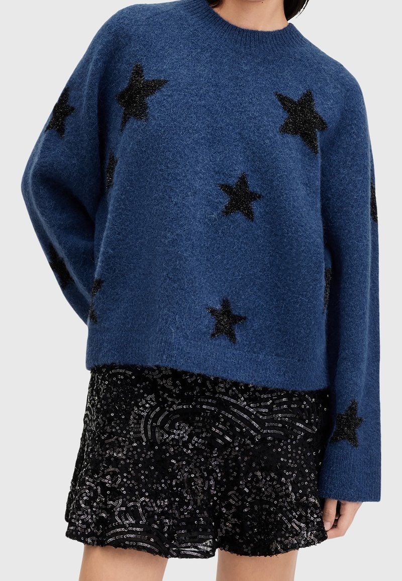 Pull bleu à texture duveteuse avec des étoiles noires, assorti d'une jupe noire à paillettes, porté par une personne avec la main reposant le long du corps.