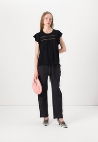 Black, polka-dotted blouse with ruffled sleeves, paired with loose, black pants. Holds a round, soft pink bag; features metallic, pointed shoes.
