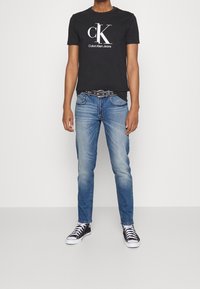 Camiseta de algodón negra con el logo blanco de Calvin Klein, combinada con jeans de denim de color azul claro y zapatillas en blanco y negro. Diseño de atuendo casual.