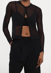 Černý průsvitný kardigan s dlouhými rukávy, kulatým výstřihem, zapínáním na knoflíky vpředu a přizpůsobeným střihem. Kombinováno s černým topem a volnými černými kalhotami.