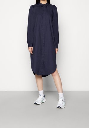 Kvinde iført en marineblå langærmet skjortekjole med hvide sneakers og hvide sokker, stående mod en ensfarvet hvid baggrund.