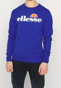Sweat-shirt bleu royal à manches longues, avec poignets et ourlet côtelés, arborant un logo "ellesse" blanc et un emblème coloré au-dessus.