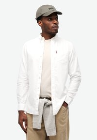 Camisa blanca de botones sobre una camiseta ligera beige, suéter gris atado a la cintura, pantalones caqui y una gorra gris con logo.