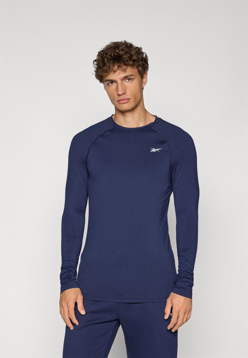 Cămașă sport cu mânecă lungă de culoare navy, realizată din material elastic, prezentând un guler rotund și un logo subtil pe piept. Textură netedă, design ajustat.