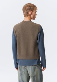 Jovem vestido com um colete de malha castanho sobre uma camisa azul de mangas longas e jeans azuis desbotados, virado de lado contra um fundo branco liso.