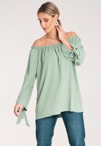 Jasnozielona bluzka na ramiączkach z marszczonym dekoltem i długimi rękawami, z ozdobnymi wiązaniami przy mankietach. Gładka faktura materiału.