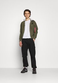 Olivgrüne Bomberjacke mit grauer Kapuze, weißem T-Shirt, schwarzen Cargo-Hosen und schwarzen Sneakern mit roten und weißen Akzenten.