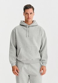 Mężczyzna w jasnoszarym hoodie i pasujących dresach, stojący na jednolitym białym tle, zwrócony przodem z neutralnym wyrazem twarzy.