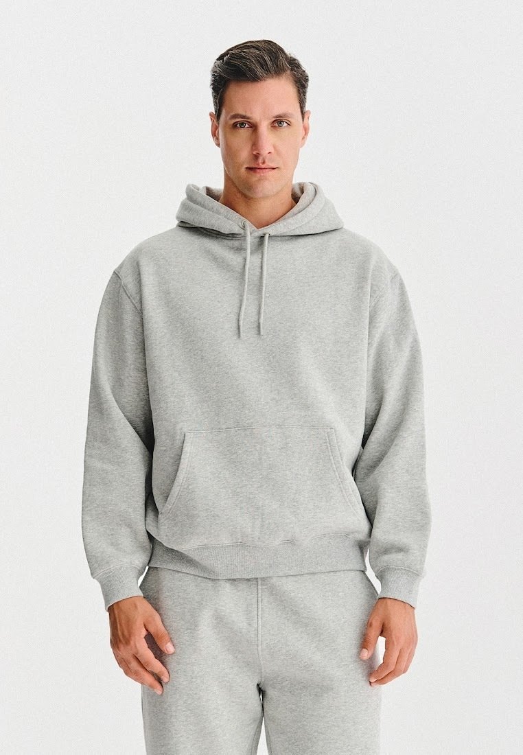 Mężczyzna w jasnoszarym hoodie i pasujących dresach, stojący na jednolitym białym tle, zwrócony przodem z neutralnym wyrazem twarzy.