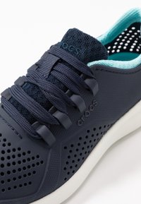 Chaussure de sport bleu marine avec dessus en mesh et caoutchouc, design perforé, semelle blanche et lacets bleu foncé. Marque visible sur le côté.