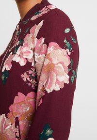 Giubbotto bomber bordeaux scuro con stampa floreale in rosa, crema e verde. Presenta un colletto a costine e chiusura con zip. Tessuto morbido al tatto.