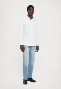 Hvit langermet skjorte med krage som kan kneppes ned, brukt sammen med lyseblå rette jeans og svarte sko på en nøytral bakgrunn.