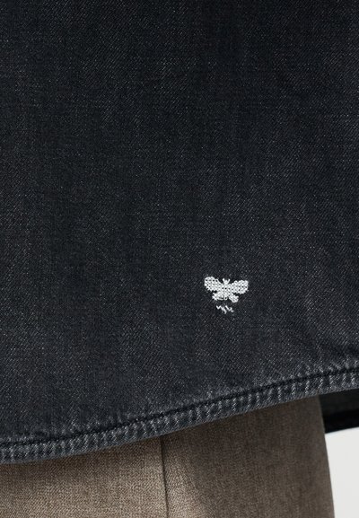 Μαύρο τζιν ύφασμα με διακριτική υφή, με ένα μικρό λευκό κεντητό λογότυπο μέλισσας κοντά στην άκρη, και καθαρό, ευθύ κόψιμο.