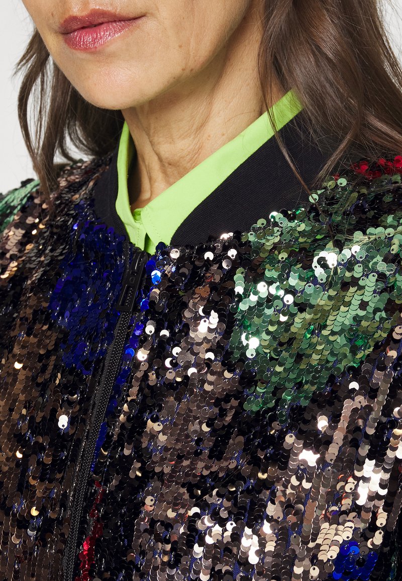 Veste bomber en sequins avec un mélange de sequins verts, bleus, dorés et noirs, dotée d'une fermeture éclair à l'avant et d'un col noir contrastant, portée sur une chemise vert citron.