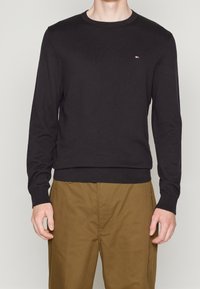Maglione nero a maniche lunghe realizzato in un tessuto morbido; presenta un collo rotondo e un piccolo logo sul petto; polsini e orlo a coste.