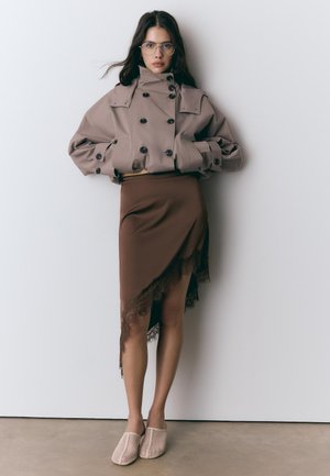 Femme portant une veste beige courte à double boutonnage, une jupe asymétrique marron ornée de dentelle, des chaussures beiges en mesh sans lacets et des lunettes, debout contre un mur blanc.