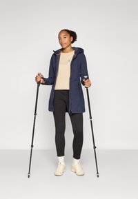 Mörkblå lång jacka med huva och kontrasterande röda detaljer, bärs över en beige topp och svarta leggings; håller i trekkingstavar; beige skor.