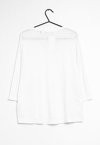 Valkoinen pitkähiha shirt, joka roikkuu mustassa vaateripustimessa valkoista taustaa vasten, näkymä takaa.