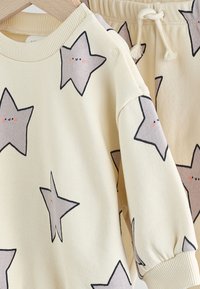 Ensemble sweat-shirt et pantalon de couleur crème avec motifs d'étoiles grises, poignets côtelés et pressions aux épaules pour un habillage facile.