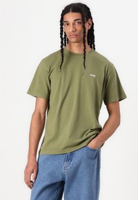 Olive green cotton t-shirt with short sleeves featuring a small "obey" logo. Paired with light blue denim jeans. Simple, casual style.