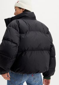 Black puffer jacket with a high collar, zippered closure, and padded sections. Features a ribbed green cuff and back pocket.