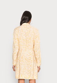 Vestido padrão amarelo e branco com mangas longas, cintura ajustada e colarinho. Apresenta um corte solto e uma textura de tecido suave.