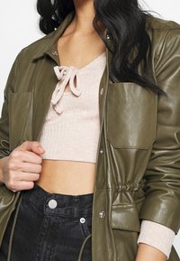 Veste en cuir vert olive avec boutons-pression, deux poches plaquées et une taille ajustable. Associée à un crop top côtelé rose clair.