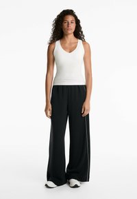 Débardeur blanc à côtes avec décolleté en V associé à un pantalon noir ample avec des rayures blanches sur les côtés et des chaussures de sport.