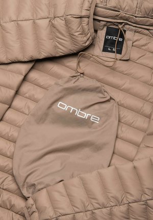 Giacca trapuntata beige con un'etichetta attaccata. Include una sacca con cordino abbinata. Il materiale è morbido e leggero, con una leggera lucentezza.