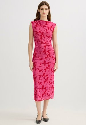 Joven mujer de pie, que lleva un vestido rosa sin mangas de largo medio con estampado floral y zapatos negros de punta puntiaguda sobre fondo neutro.