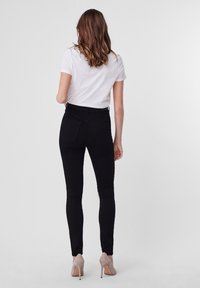 Jean skinny noir taille haute avec poches arrière, associé à un t-shirt blanc uni et des talons hauts beiges, vu de dos.