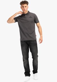 Šedé polo tričko s límečkem a třemi knoflíky, logo na hrudi, spárované s tmavými džíny a bílými teniskami, stojící v neutrální pozici.