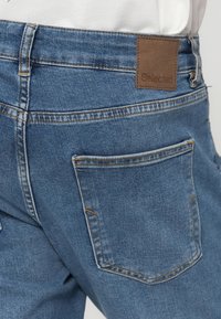 Zilas džinsa bikses ar klasisku piegriezumu, kurām ir brūns ādas zīmola uzraksts, aizmugurējā kabata un kontrastējošas šūšanas detaļas.