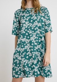 Vestido verde azulado hecho de una tela suave con un corte holgado, que presenta un patrón floral blanco. Mangas cortas y un escote redondo.