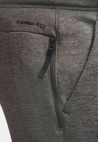 Gros plan sur un pantalon en tissu gris avec une poche zippée noire verticale et l'inscription "THERMAL-FITT" brodée au-dessus de la poche.