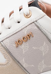 Baskets avec cuir blanc et gris, panneaux texturés, détail du logo doré, lacets blancs plats et un motif floral subtil sur un côté.