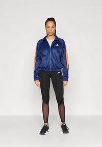 Tamsiai mėlynas striukės su užtrauktuku, oranžinėmis rankovėmis; juodos tamprės su tinklelio detalėmis; juodi sportiniai batai. Ant drabužių yra „Adidas“ logotipas.