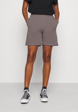 Persona con pantalones cortos grises informales, camiseta negra y zapatillas negras altas con cordones blancos, de pie con las manos en los bolsillos.