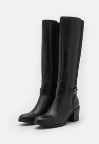 Botas de cuero negras hasta la rodilla con tacones cuadrados, cremalleras laterales y correas decorativas con pequeñas hebillas metálicas sobre un fondo blanco.