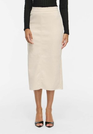 Kvinde iført beige midi-nederdel med højt talje, sort textureret langærmet top og sorte åbne hæle, stående på hvid baggrund.