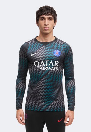 Άνδρας φοράει φανέλα ποδοσφαίρου της Paris Saint-Germain με μαύρο, μπλε και γκρι ζιγκ-ζαγκ μοτίβο και λογότυπο Qatar Airways, στέκεται μπροστά σε απλό φόντο.