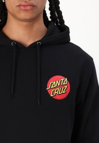 Μαύρη κουκούλα από βαμβακερό μείγμα, με κυκλικό κόκκινο λογότυπο που έχει την ένδειξη "SANTA CRUZ" σε κίτρινες γράμματα στο αριστερό στήθος. Περιλαμβάνονται κορδόνια.