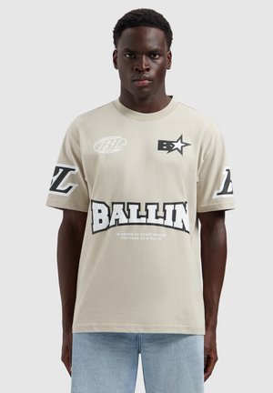 Man draagt een beige T-shirt met zwart-witte "BALLIN" tekst en logo's, gecombineerd met lichtblauwe spijkerbroek, staand tegen een lichte achtergrond.