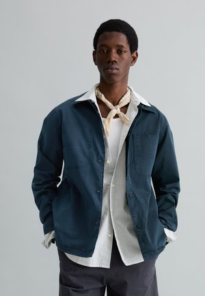 Młody mężczyzna ubrany warstwowo: niebieska kurtka, pasiasta koszula, biały podkoszulek, beżowy szalik na szyi oraz ciemne spodnie na jednolitym tle.