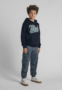 Sweat à capuche noir avec un motif "Utah", pantalon cargo gris avec poches, et baskets blanches avec semelles beiges. Look décontracté et matériaux confortables.